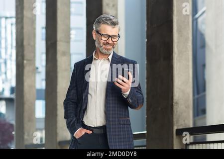 Successful gray haired boss walking outside office building, mature businessman using phone, smiling and reading news typing message, gray haired man in business suit and glasses. Stock Photo
