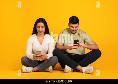 Cheerful busy millennial arab guy and lady in casual sit on floor, typing on smartphone, searching, surfing in internet Stock Photo