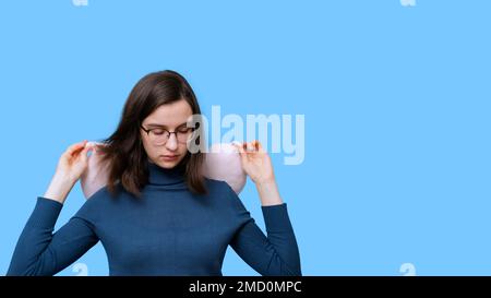 Portrait of a student girl in glasses and a sweater sleeping in a travel pillow on a blue background, banner, copy space. A woman at rest with a pillo Stock Photo