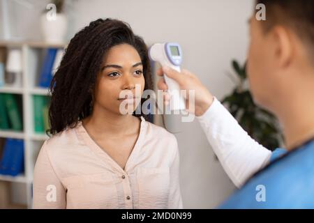 Serious middle aged korean doctor taking temperature with non-contact thermometer of black millennial female Stock Photo