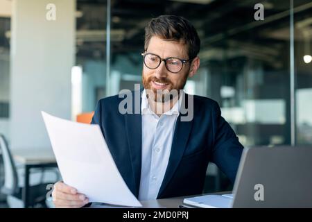 Busy middle aged businessman working with laptop and documents, sitting at desk in office, checking financial reports Stock Photo