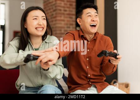 Excited asian couple playing video games at home, happy asian emotional man and woman sitting on couch with joysticks Stock Photo