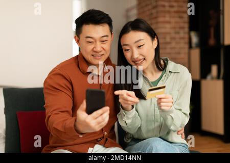 Happy asian man and woman holding smartphone and credit card, shopping online from home, sitting on sofa Stock Photo