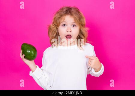 Kid hold red avocado in studio. Studio portrait of cute child with avocado isolated on pink background. Stock Photo