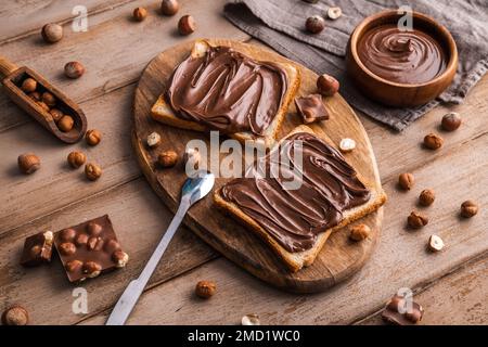 Board of bread with chocolate paste and hazelnuts on wooden background. Popular desser food. Top view. Stock Photo