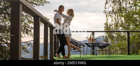 Horizontal sideview shot of a young couple in summer outfit enjoying the mountain view from terrace. Copy space. Stock Photo