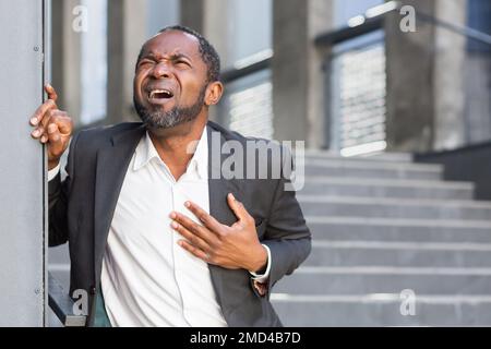 Heart attack, African American man has severe chest pain, businessman outside office building on stairs writhing in chest pain. Stock Photo
