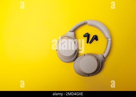 Light gray wireless on-ear headphones and blue in-ear wireless headphones on a yellow background. Comparison of different types of headphones. Headpho Stock Photo