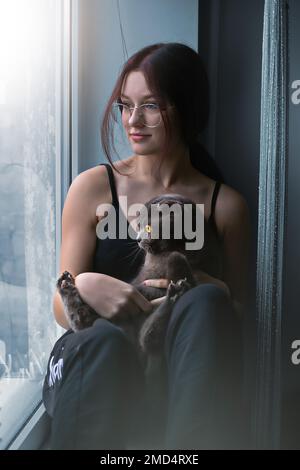 Attractive teenage girl sits alone on the windowsill together with a pet python with a cat and looks out the window. High quality photo Stock Photo