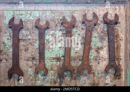 Five old, rusted wrench tools hanging on nails on cracked, damaged wall. Stock Photo