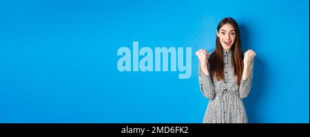 Excited woman looking relieved and hopeful, making fist pump and smiling, winning prize, celebrating achievement or victory, standing on blue Stock Photo