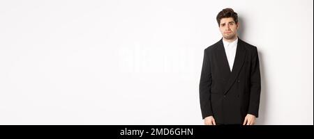 Image of gloomy and sad handsome male model in black suit, frowning and looking tired at camera, standing against white background Stock Photo
