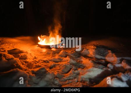 A fire burns in a fire pit at a campsite in winter. Snow surrounds the metal container where orange flames light up the surrounding snow in the dark. Stock Photo
