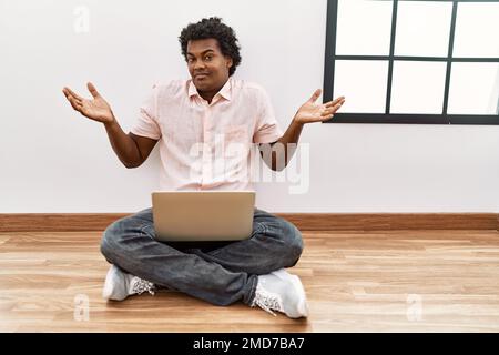 African man with curly hair using laptop sitting on the floor clueless and confused expression with arms and hands raised. doubt concept. Stock Photo