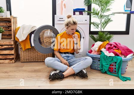 Young blonde woman doing laundry sitting by washing machine feeling unwell and coughing as symptom for cold or bronchitis. health care concept. Stock Photo