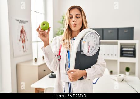 Young blonde doctor woman holding weighing machine and green apple making fish face with mouth and squinting eyes, crazy and comical. Stock Photo