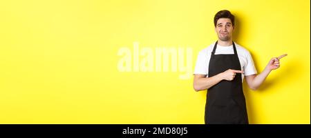 Doubtful barista in black apron pointing fingers right, looking skeptical and unamused, standing over yellow background Stock Photo