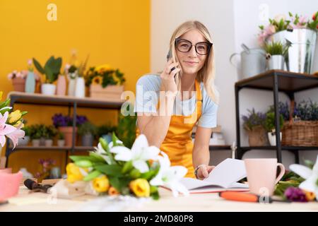 Young blonde woman florist talking on smartphone reading book at flower shop Stock Photo