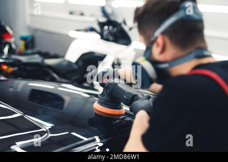 Professional car detailer polishing black car paintwork with orbital polisher. Back view. Blurred silhouette in the foreground. High quality photo Stock Photo