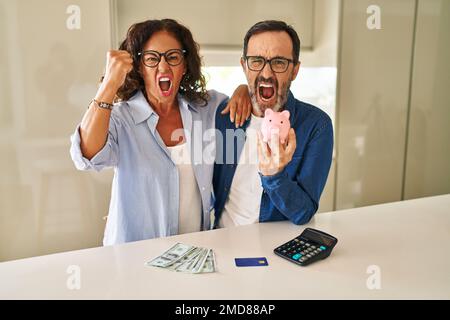 Middle age couple holding piggy bank calculating savings annoyed and frustrated shouting with anger, yelling crazy with anger and hand raised Stock Photo