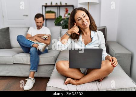 Hispanic middle age couple at home, woman using laptop smiling doing phone gesture with hand and fingers like talking on the telephone. communicating Stock Photo