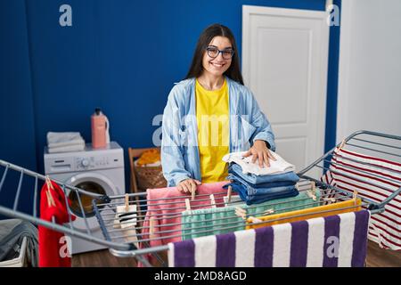 Young hispanic woman holding folded jeans hanging clothes on clothesline at laundry room Stock Photo