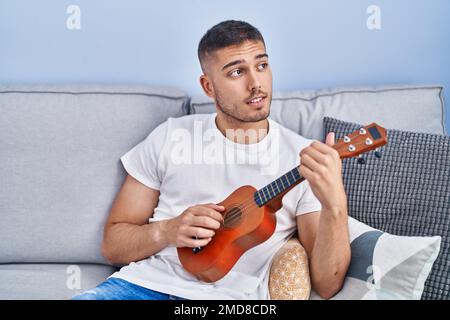 Young hispanic man playing ukelele sitting on sofa at home Stock Photo