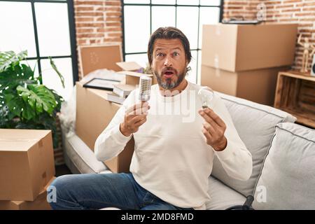 Handsome middle age man holding led lightbulb and incandescent bulb in shock face, looking skeptical and sarcastic, surprised with open mouth Stock Photo