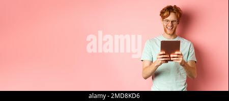 Happy guy with red hair and beard using digital tablet, reading screen and smiling, standing over pink background Stock Photo