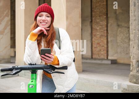 Smiling young redhead woman with mobile phone, rents e-scooter using smartphone application, riding around city, urban transport and people concept Stock Photo