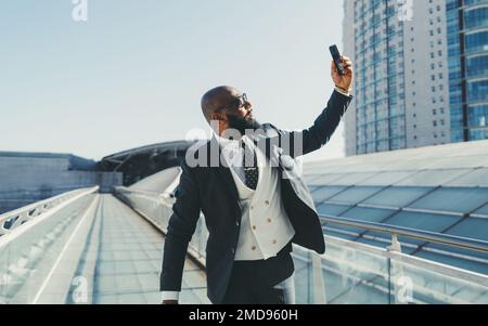 A capture of a black, bald, and unshaven businessman, looking gallant and damper in his elegant full-body suit taking a selfie on a glassed-in, elevat Stock Photo