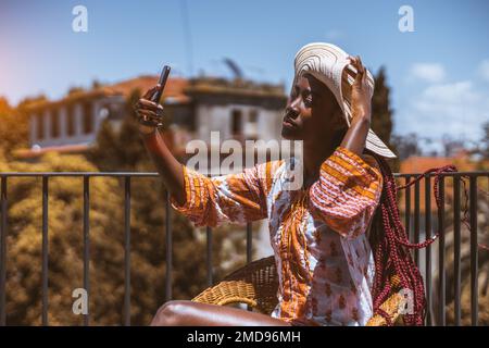 A young woman, with pink long box braids, wearing a colorful short dress with long sleeves, and a wide-brimmed hat, taking a selfie on a rooftop fence Stock Photo