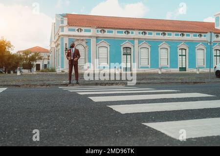 A bald black man looking dapper in his Bordeaux custom-made suit standing in front of the zebrawalk waiting for the green light to cross the road, in Stock Photo