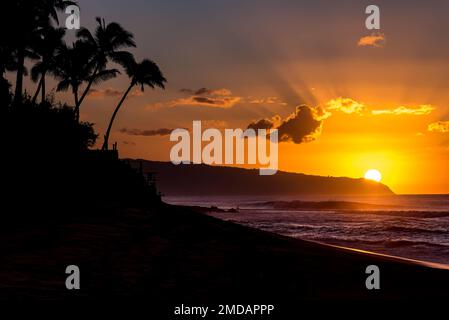 Sun setting behind the mountain over waves and palm trees on Sunset Beach, Hawaii Stock Photo