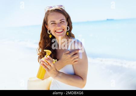Portrait of smiling stylish 40 years old woman at the beach in white beachwear applying spf. Stock Photo