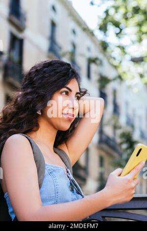 vertical portrait of an attractive young indian woman with yellow smart phone on her hands. She is leaning on a fence and looking away Stock Photo