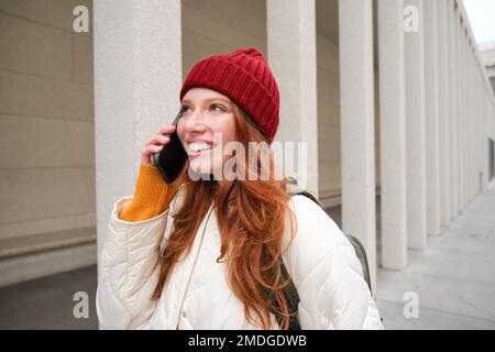 Mobile broadband and people. Smiling young redhead woman walks in town and talks on mobile phone, calling friend on smartphone, using internet to make Stock Photo