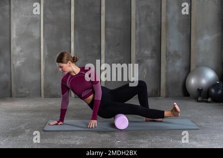 A young woman in sportswear performs a myofascial hip massage with a roller. Muscle recovery, mfr. Stock Photo