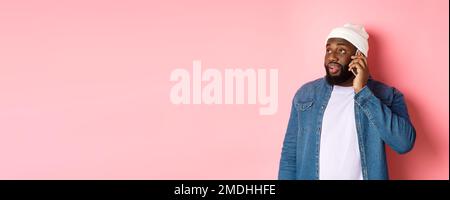 Hipster Black man talking on phone, looking left and having mobile conversation, standing over pink background Stock Photo