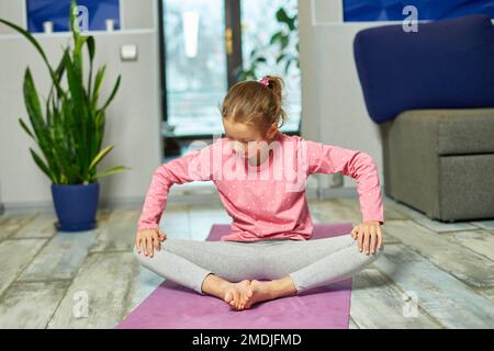 Foto de Little girl doing stretching exercises, practicing yoga on fitness  mat at home do Stock