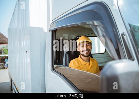 bearded delivery man in yellow uniform smiling while driving truck Stock Photo
