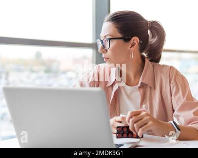 Frowning woman looks on medicine pills while working with laptop. Mental health problems, emotional burnout or headache. Modern office at co-working c Stock Photo