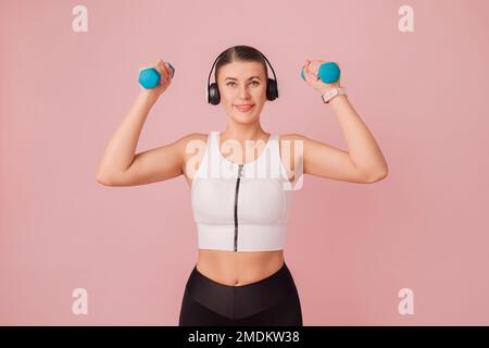 Smiling young woman in headphones doing sport exercise while standing over pastel pink backdrop. Studio portrait of cheerful fitness woman Stock Photo