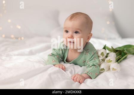 cute baby in a green cotton bodysuit at home on a white bed with tulips. Spring, Mother's Day, March 8. High quality photo Stock Photo