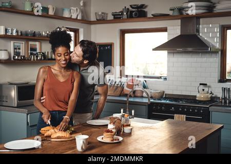 Im looking forward to take a bite. a man kissing his wife while she prepares breakfast in the kitchen at home. Stock Photo