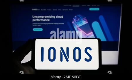 Person holding smartphone with logo of German web hosting company Ionos SE on screen in front of website. Focus on phone display.