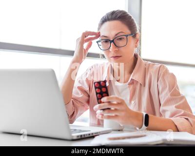 Frowning woman looks on medicine pills while working with laptop. Mental health problems, emotional burnout or headache. Modern office at co-working Stock Photo