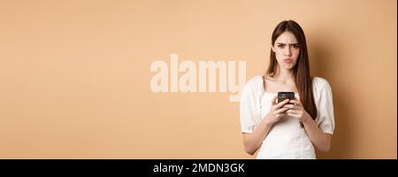 Disappointed girl with smartphone frowning, pucker lips upset, reading bad news on phone, standing on beige background Stock Photo