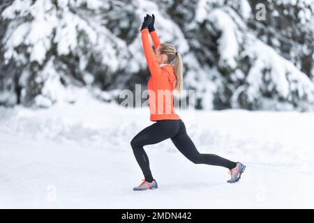 Athletic female runner doing stretching exercise, preparing for workout in the snowy winter park. Stock Photo