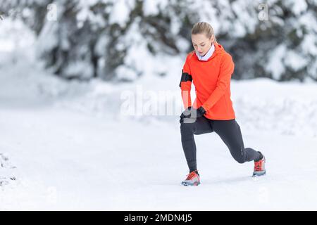 Athletic female runner doing stretching exercise, preparing for workout in the snowy winter park. Stock Photo